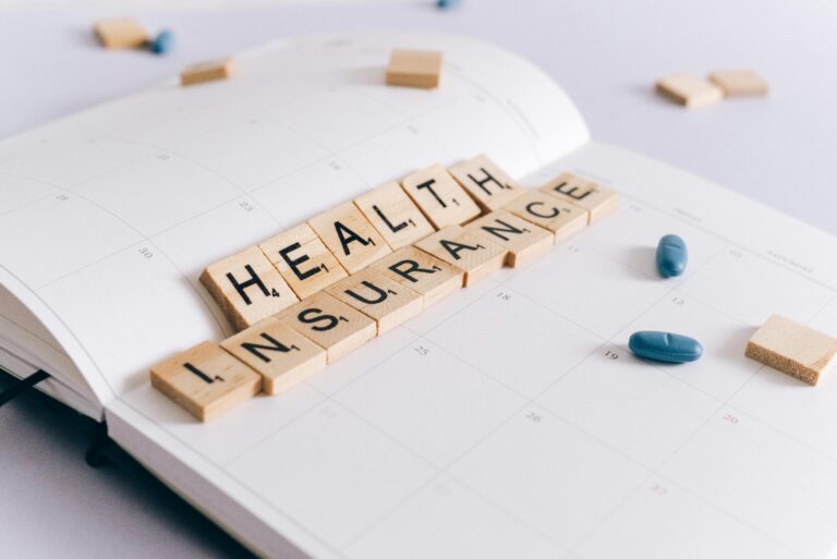 Fichas de madera que forman las palabras "HEALTH INSURANCE" sobre un calendario abierto, acompañadas de dos cápsulas azules y fichas en blanco.
