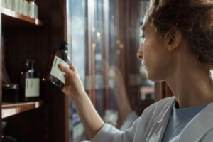 Una profesional de la salud con bata blanca observa detenidamente la etiqueta de un frasco de medicina oscuro mientras lo retira de un estante de madera.