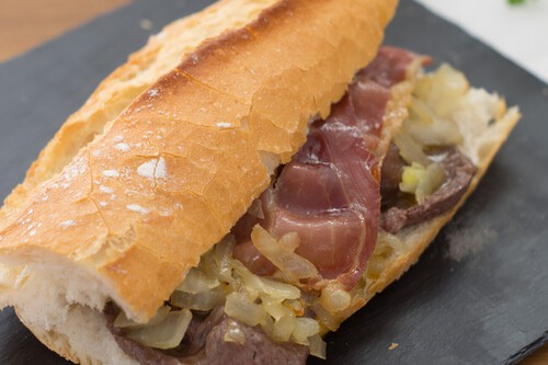 A close-up of a rustic baguette sandwich filled with grilled pork loin, serrano ham, and caramelized onions on a slate board.