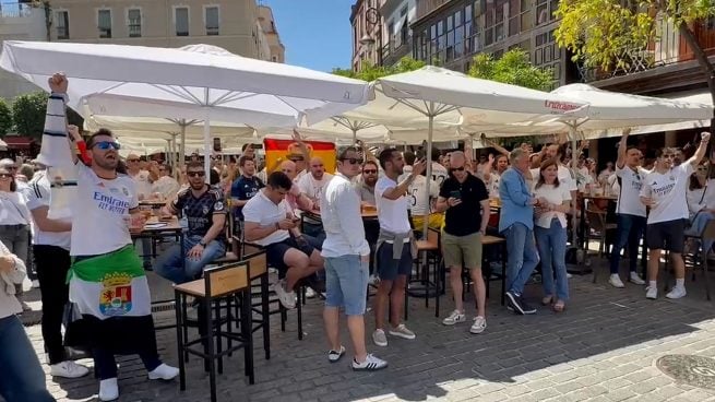 A large group of football fans in white jerseys celebrating at an outdoor terrace with white umbrellas in a sunny city square.
