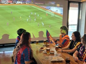 A group of friends sitting at a long wooden table in a bar, watching a football match on a large projector screen.