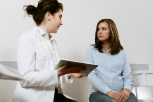 A female doctor in a white coat holding a tablet while talking to a young female patient in a light blue shirt during a medical appointment.