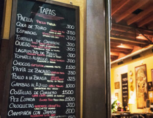 A hand-written chalkboard menu in a Spanish bar listing various traditional tapas like Paella, Cola de Toro, and Gambas al Ajillo with their prices.