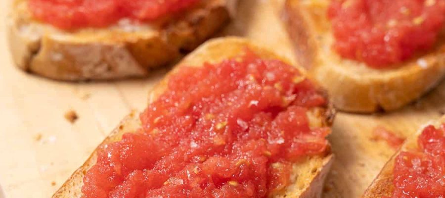Four slices of toasted rustic bread topped with fresh grated tomato and salt, served on a wooden board.