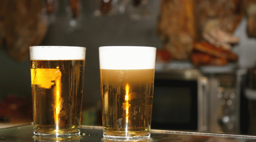 Two glasses of cold, amber-colored beer with a thick head of foam sitting on a bar counter with a blurred tavern background.