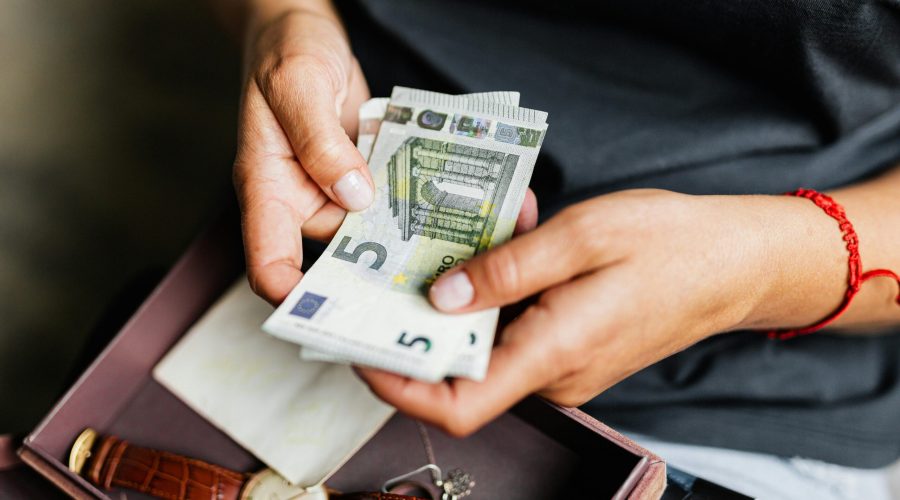 A close-up of a person's hands counting 5-euro banknotes over an open box containing a leather-strap wristwatch.