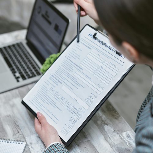 Vista desde arriba de una mujer sosteniendo un portapapeles con un formulario titulado "HOME INSURANCE POLICY" mientras señala con un bolígrafo.