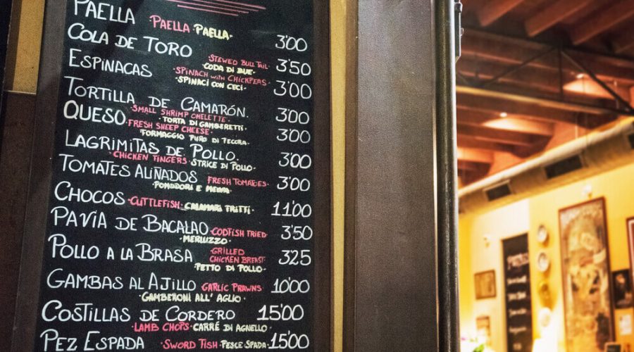 A hand-written chalkboard menu in a Spanish bar listing various traditional tapas like Paella, Cola de Toro, and Gambas al Ajillo with their prices.